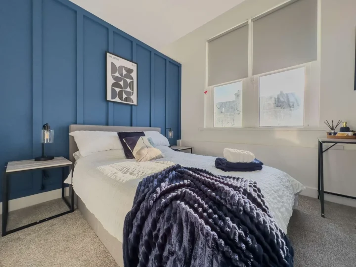 bedroom with white and blue walls with a grey bed covered with purple and white bedding, cushions and towels